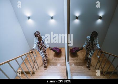 A wide-angle shot of a youthful charming black female traveler rising the stairs with her pink wheeled suitcases; a cheerful young African woman is ar Stock Photo