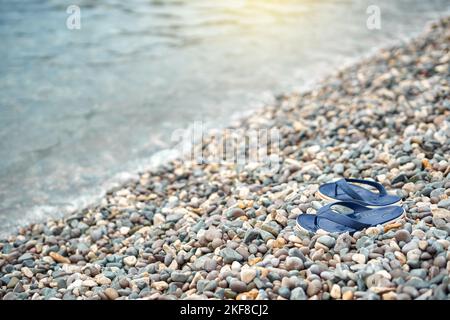 Pair of bright blue flip-flops left on pebble beach against blurry blue water Stock Photo