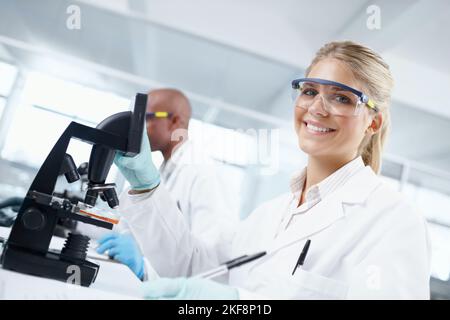 Scientists must ask the right questions in order to find the rig. Portrait of a gorgeous female scientist in front of a microscope with a male Stock Photo