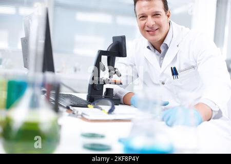 Research saves lives. A researcher smiling while sitting at a table in front of a microscope and computer. Stock Photo