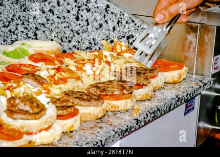 Selective focus on cooker hand, tong and burgers filled with french fries, lettuce, tomato, grilled chopped steak, mayonnaise and ketchup sauce. Stock Photo