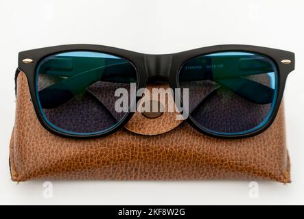 Black frame blue sun-glass made of acetate material resting on a brown leather case on white isolated background. Selective Focus in center. Stock Photo