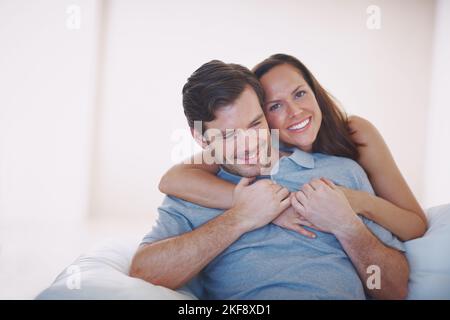 Surprising her husband with some affection. A loving young wife hugging her husband from behind while he sits on a sofa. Stock Photo