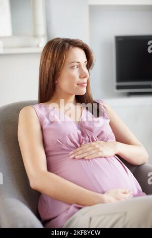 Experiencing the perfect pregnancy. Pretty young pregnant woman looking away while she sits in her living room as she holds her belly. Stock Photo