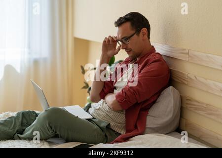Concentrated Caucasian man sits on bed in hotel room reading news via internet or checking email Stock Photo