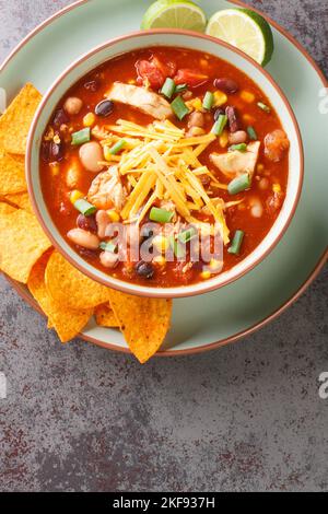 Mexican Creamy Chicken Taco Soup with black bean, corn, tomatoes, tortilla chips topped with cheddar cheese in green bowl on a table, Vertical top vie Stock Photo