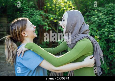 Strong female friendship. Happy two teen girls best friends holding hands and hugging while standing in front of park. Multiethnical friends Stock Photo