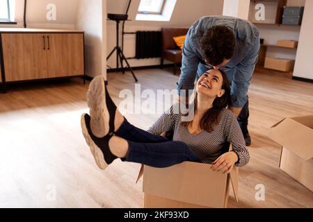Happy brunette couple having fun and riding in cardboard boxes in new home. Home,  smiling people, moving and real estate concep Stock Photo