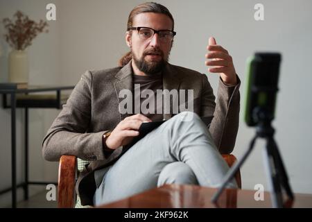 Bearded mature therapist having online consultation with patient sitting in front of smartphone on tripod and talking to patient Stock Photo