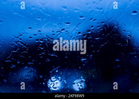 Glass with focused raindrops and blurred background, at night, in a blue tone. with the headlights of a car from the front. Concept of rain, winter, s Stock Photo