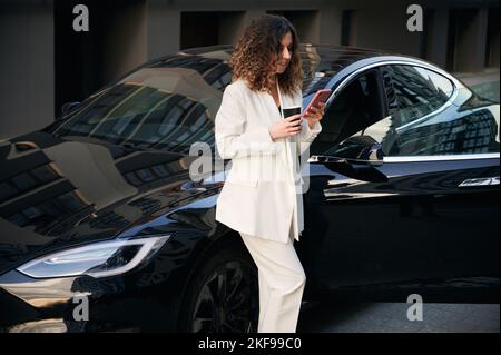 Stylish businesswoman holding cup of coffee and using cellphone while standing by electric car on the street. Elegant young woman reading message on mobile phone while leaning on black automobile. Stock Photo