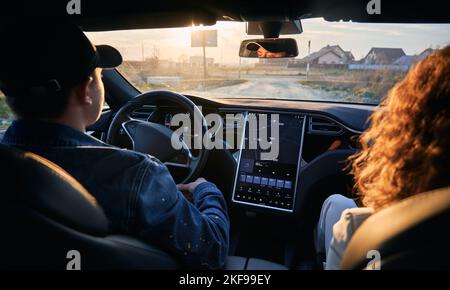 Rear view inside the car. Driver and passenger travelling by electric car with modern user interface with touch screen on dirt road. Male chauffeur riding on terrain using navigation system. Stock Photo