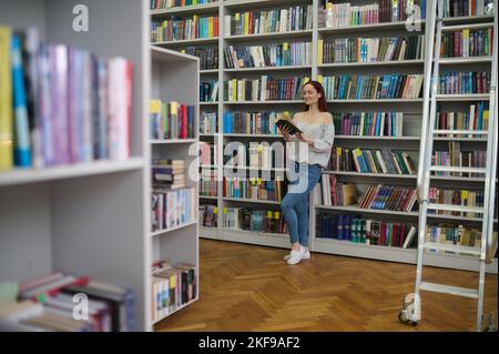 Young librarian standing near the bookshelves with a gadget in hands Stock Photo