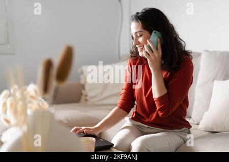 Young happy woman talking on the phone and typing on the computer sitting on the couch at home Stock Photo