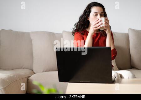 Young woman having a coffee and looking sideways the computer screen sitting on the couch at home. Working from home concept Stock Photo