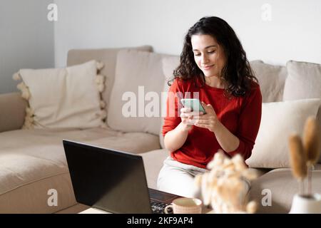 Young happy woman using the phone and  the computer sitting on the couch at home Stock Photo