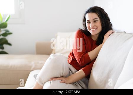 Young beautiful brunette woman smiling to camera while resting in the couch at home Stock Photo