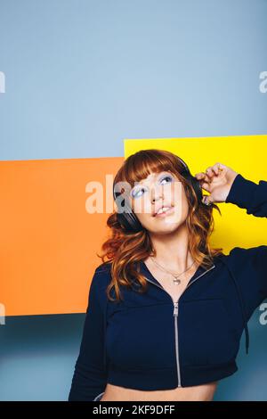 Thoughtful woman listening to her headphones with graphic shapes in the background. Pensive young woman enjoying an interesting audio playlist in a st Stock Photo