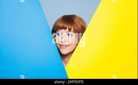 Smiling woman with ginger hair and blue eyeshadow looking away while standing behind colour placards. Happy young woman peeping from behind two advert Stock Photo