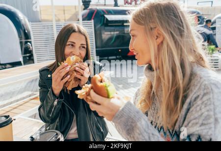Two attractive European women eat street fast food while sitting at a table outside. Stock Photo