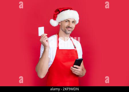mature man in christmas santa hat and red apron hold smartphone and card. advertisement Stock Photo