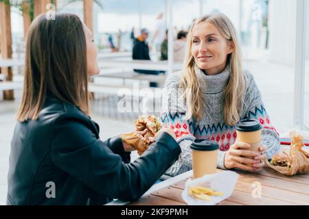 Two girlfriends eat burgers, drink coffee and chat on the street, sitting at the table. Stock Photo