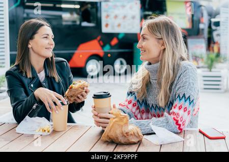 Two female friends eat hamburgers, drink coffee and chat on the street, sitting at the table. Stock Photo