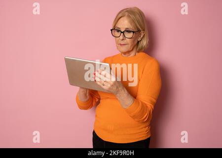 Portrait of smiling beautiful senior aged mature woman using tablet computer, isolated over pink background. Copy space for text. Stock Photo