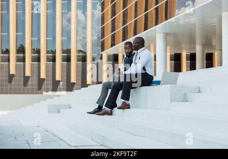 Two African American businessmen in suits communicate. Friends and business partners sit on the concrete steps of the city stairs against the backdrop Stock Photo