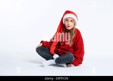 Extremely excited surprised little girl wearing santa claus hat, sitting on floor. white background Stock Photo