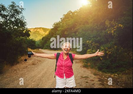 Excited happy senior woman backpacker tourist walking in summer forest road outdoors at sunset time. Old slim lady traveling with photo camera. Active retirement vacation concept. Warm filter Stock Photo