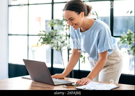 Positive concentrated busy mixed race female employee, successful business lady, working with laptop, typing text, answering email, while standing in modern office near work desk, smiling Stock Photo