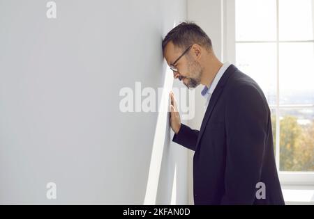Businessman gets tired of problems, feels frustrated and bangs his head against office wall Stock Photo