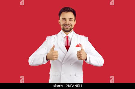 Happy young man in white suit isolated on red background smiling and showing thumbs up Stock Photo