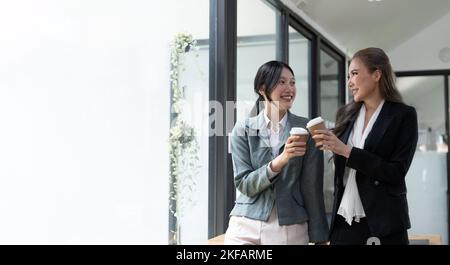 Diverse employees chatting during coffee break, walking in modern office, Asian businesswoman wearing glasses sharing ideas, discussing project with Stock Photo