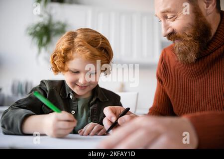 smiling redhead kid writing in notebook while doing homework near pleased dad Stock Photo