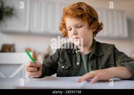 pensive redhead boy holding pen while doing homework near blurred notebook Stock Photo