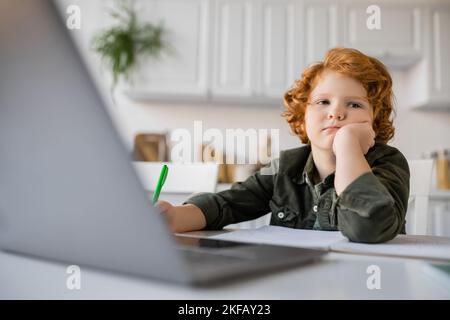 bored redhead boy sitting with hand near face while doing homework at blurred laptop Stock Photo
