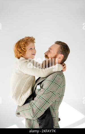 Trendy father in jacket hugging and lifting cheerful redhead son on grey background Stock Photo