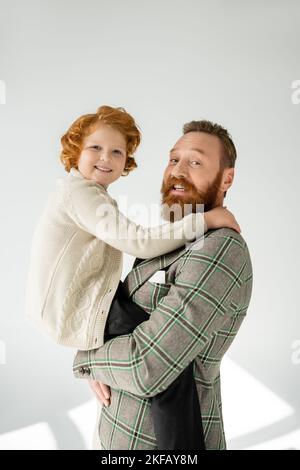 Smiling man in checkered jacket holding red haired son on grey background with sunlight Stock Photo