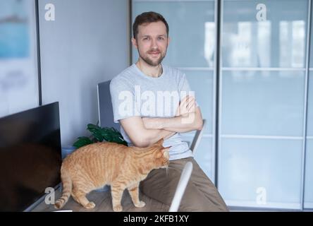 Young engineer man in gray t-shirt. Portrait of cheerful bearded caucasian male freelance software developer at working place in home office near computer with tabby cat looking at camera Stock Photo