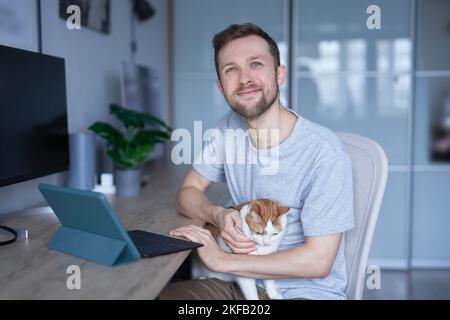 Attractive caucasian bearded young male sitting at home office with his cat using tablet for work or education. Smart working and smart studying with pet. High quality photo Stock Photo