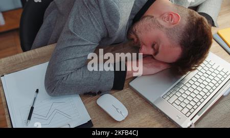 Bored sleepy businessman programmer software developer in formal wear suit falling asleep after routine work on laptop computer at office desk. Exhausted tired freelancer workaholic man. Overworked Stock Photo