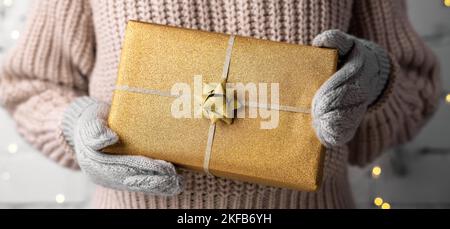 woman holding wrapped golden christmas gift in hands with wool mittens. banner Stock Photo