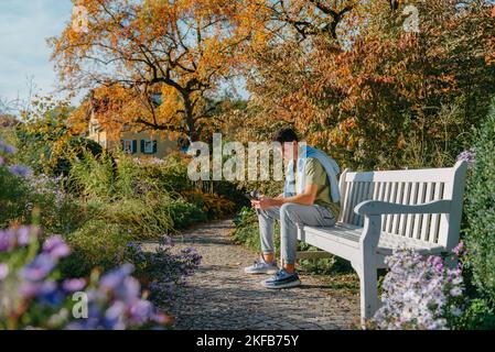 A Teenager Sits On A Bench In The Autumn Park Drinks Coffee From A Thermo Mug And Looks Into A Phone. Portrait Of Handsome Cheerful Guy Sitting On Stock Photo