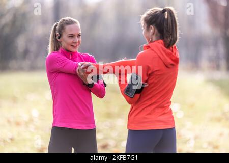 Two smiling female friends are warming up before jogging, doing upper body stretching in the park. Stock Photo