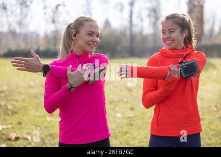Two female athletes are exercising before jogging, doing upper body stretching in the park. Stock Photo