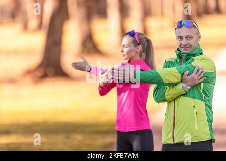 Two amateur athletes mature man and young woman are warming up before jogging, doing upper body stretching in the park.