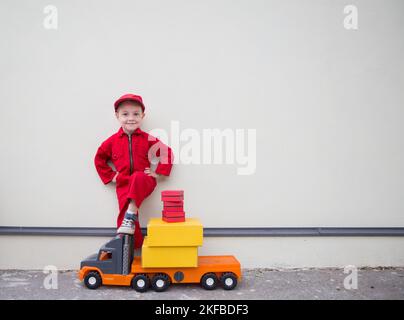 cute smiling boy 3-4 years old in red overalls and a cap stands near a big toy truck loaded with cardboard boxes. postman's little helper, truck drive Stock Photo