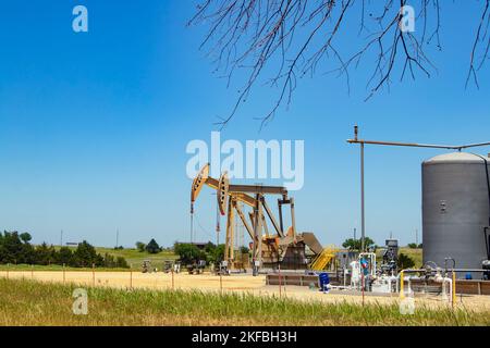 Two pump jacks at a pumping station with tanks and machinery with some farm buildings on the horizon under a blue sky with a few tree limbs hanging do Stock Photo